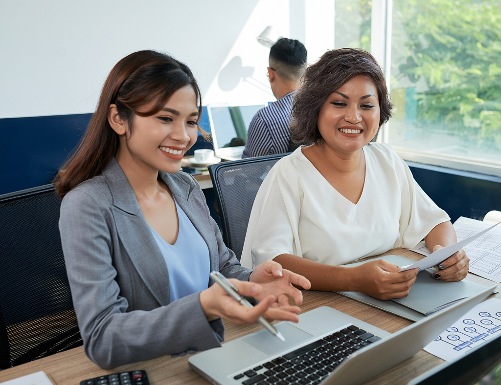 Two Asian female colleagues at a desk with a laptop, one helping the other